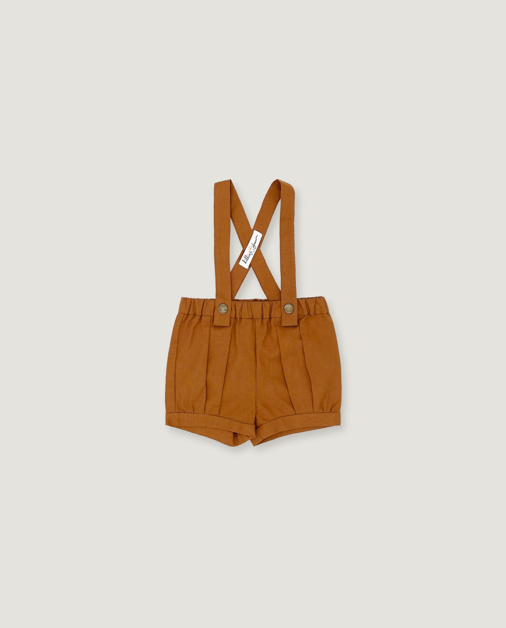 A pair of toddler's pleated shorts in a warm camel brown color, featuring adjustable shoulder straps with a crossed design and a small white brand tag.