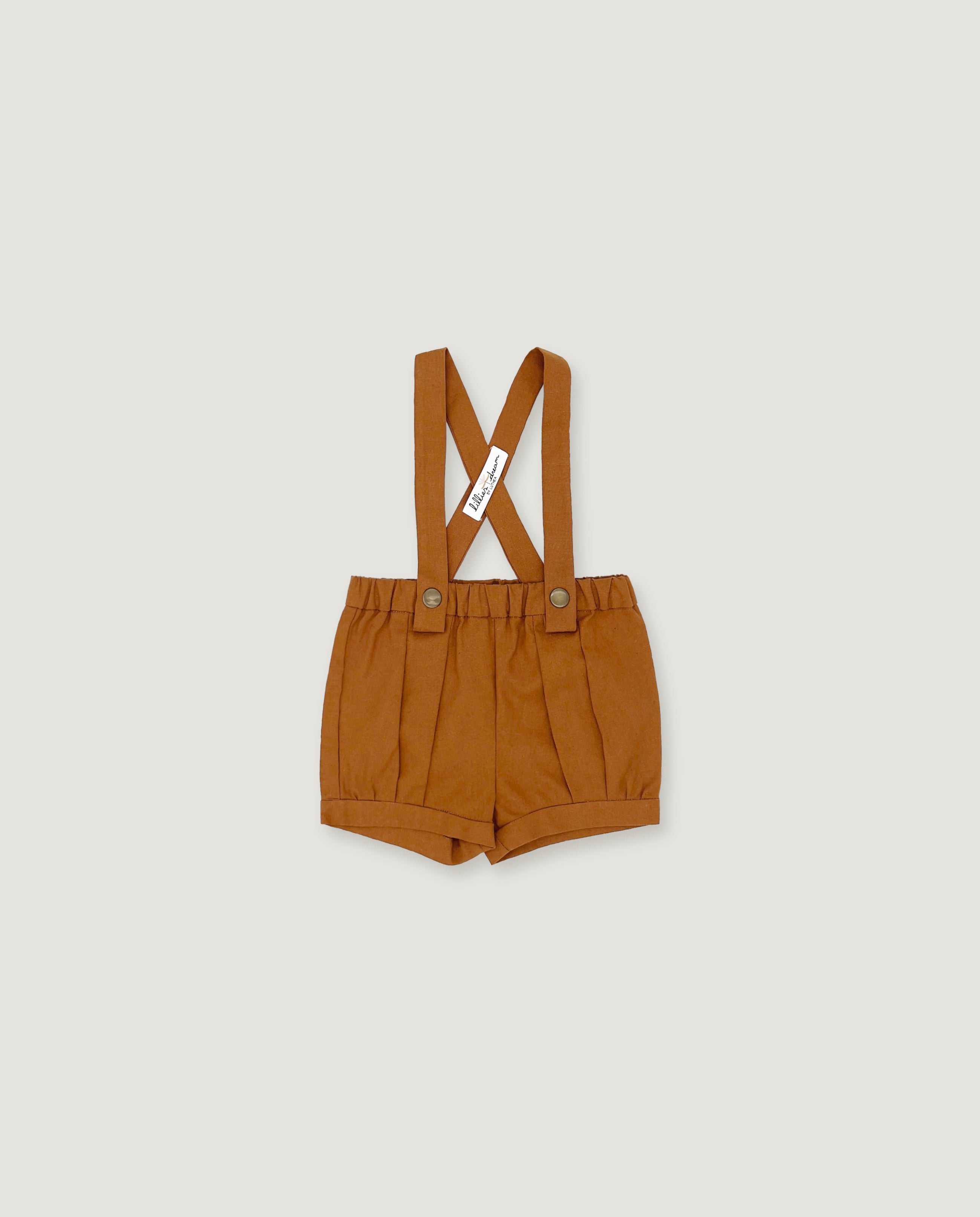A pair of toddler's pleated shorts in a warm camel brown color, featuring adjustable shoulder straps with a crossed design and a small white brand tag.