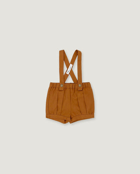 A pair of toddler's pleated shorts in a warm camel brown color, featuring adjustable shoulder straps with a crossed design and a small white brand tag.