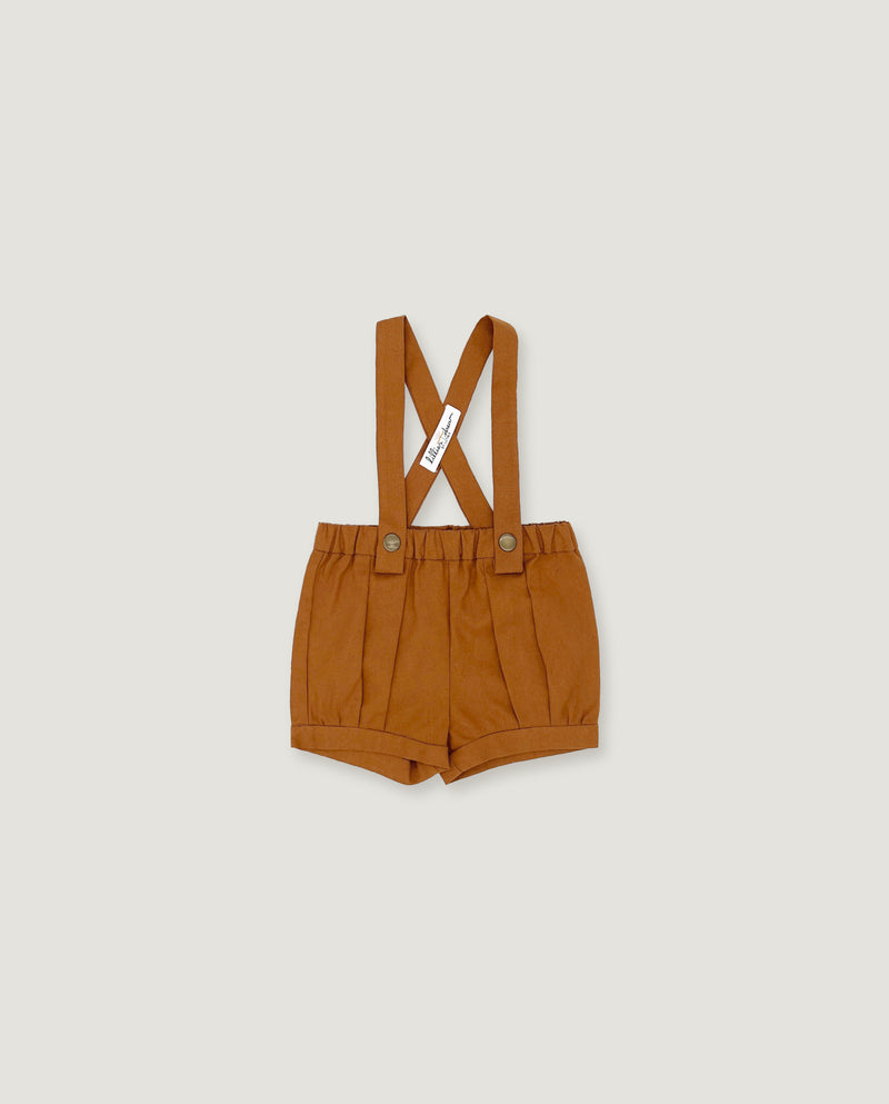 A pair of toddler's pleated shorts in a warm camel brown color, featuring adjustable shoulder straps with a crossed design and a small white brand tag.
