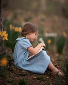 A young girl with light brown hair sits on the ground in a field of yellow daffodils, wearing a light blue and white gingham dress and holding a book.