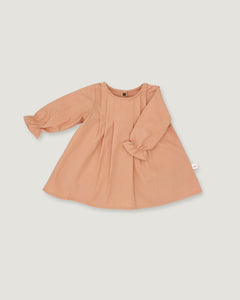 A long-sleeve baby dress in a soft peach color, featuring gathered ruffle details on the sleeves and a round neckline, laid flat against a light gray background.