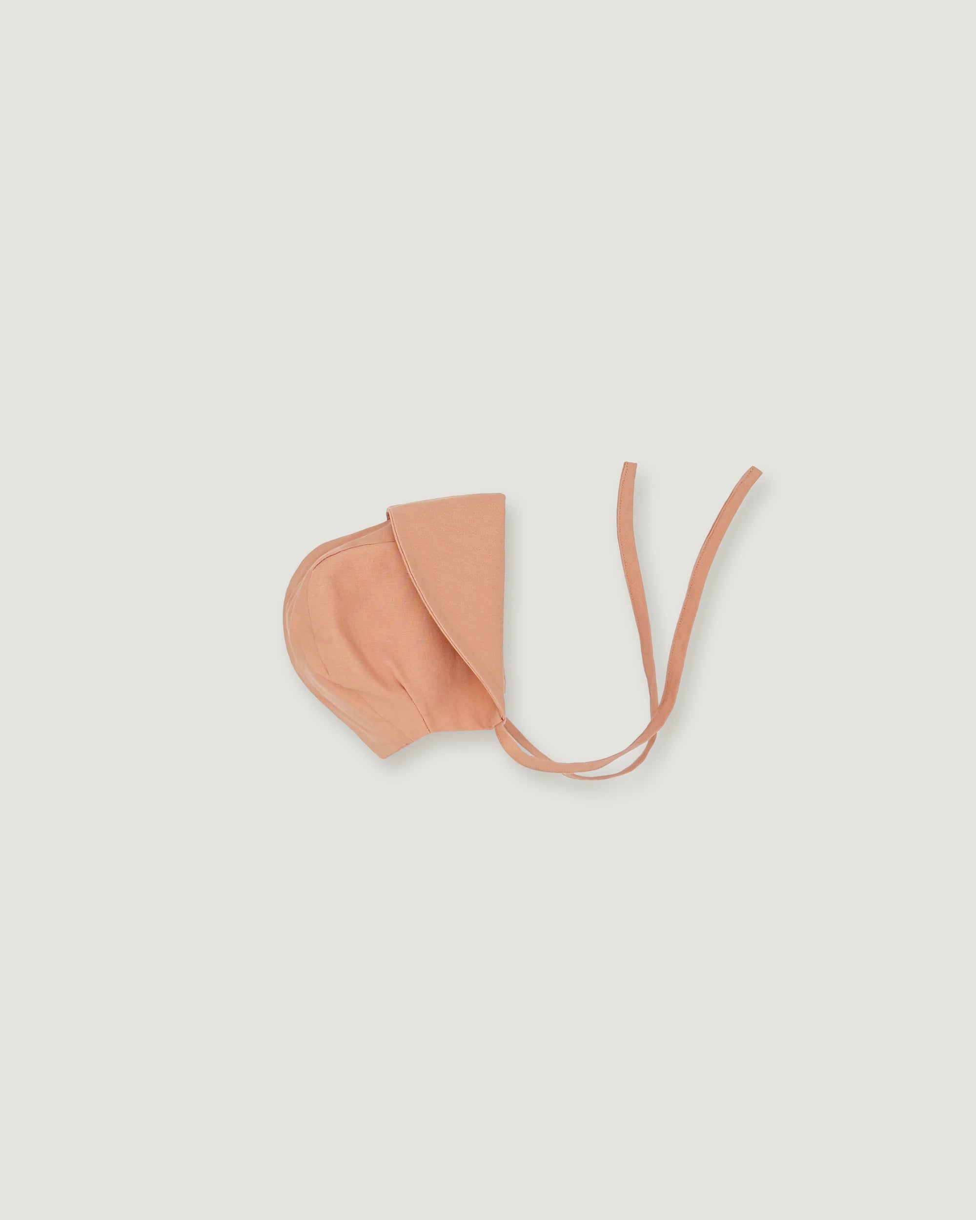 A light peach-colored fabric bonnet with long tie straps, laid flat on a solid light gray background.