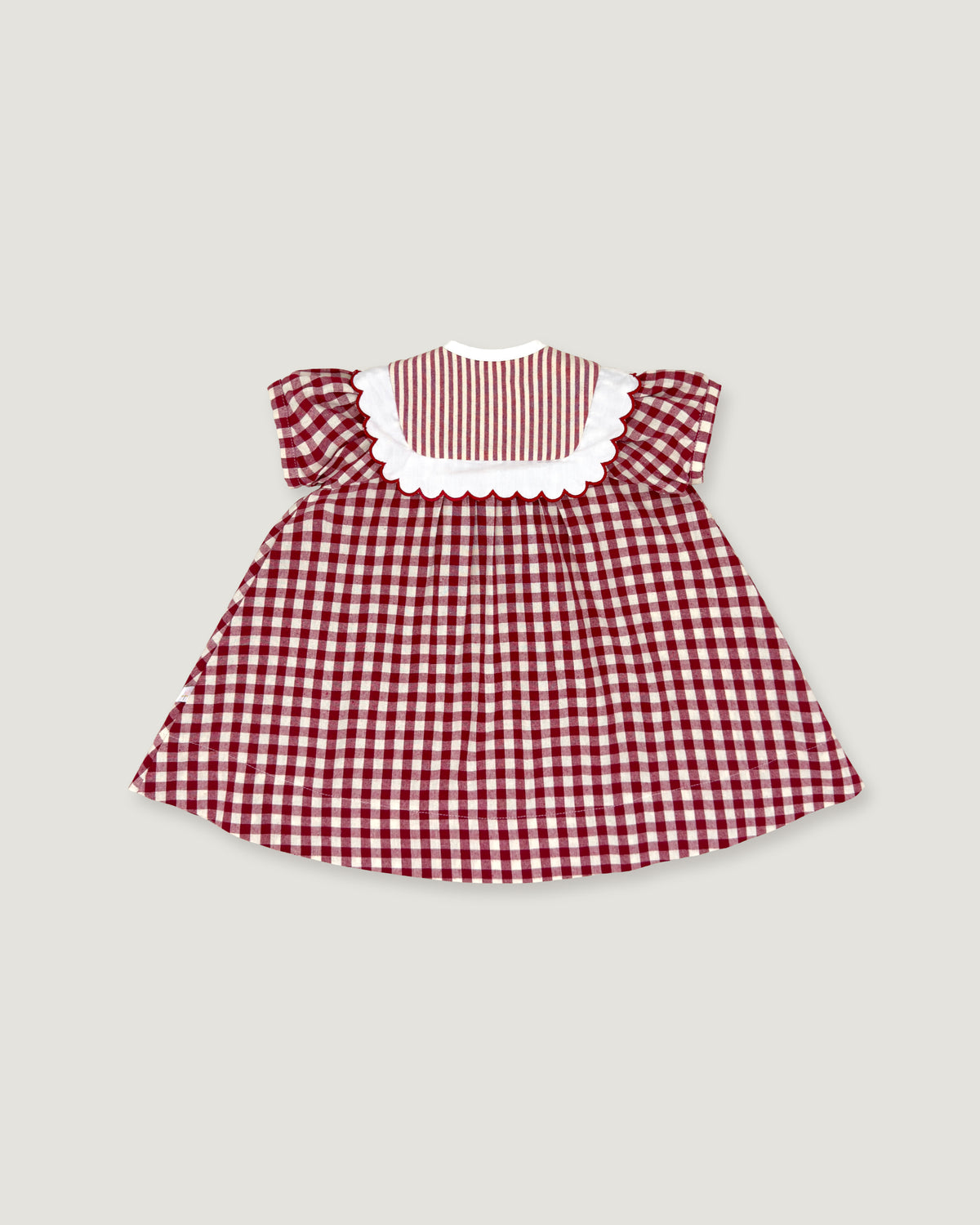 A small children's dress with a red and white gingham pattern, featuring a solid red and white striped yoke with a white scalloped collar, set against a solid light gray background.