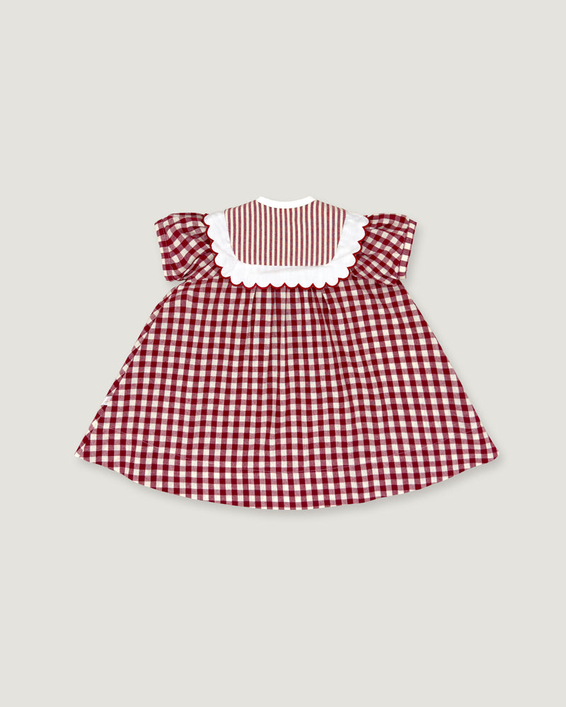 A small children's dress with a red and white gingham pattern, featuring a solid red and white striped yoke with a white scalloped collar, set against a solid light gray background.