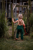 Back view of a 3-year-old girl closing a wooden gate in evergreen linen long overalls, highlighting the adjustable straps and overall functionality for everyday wear.