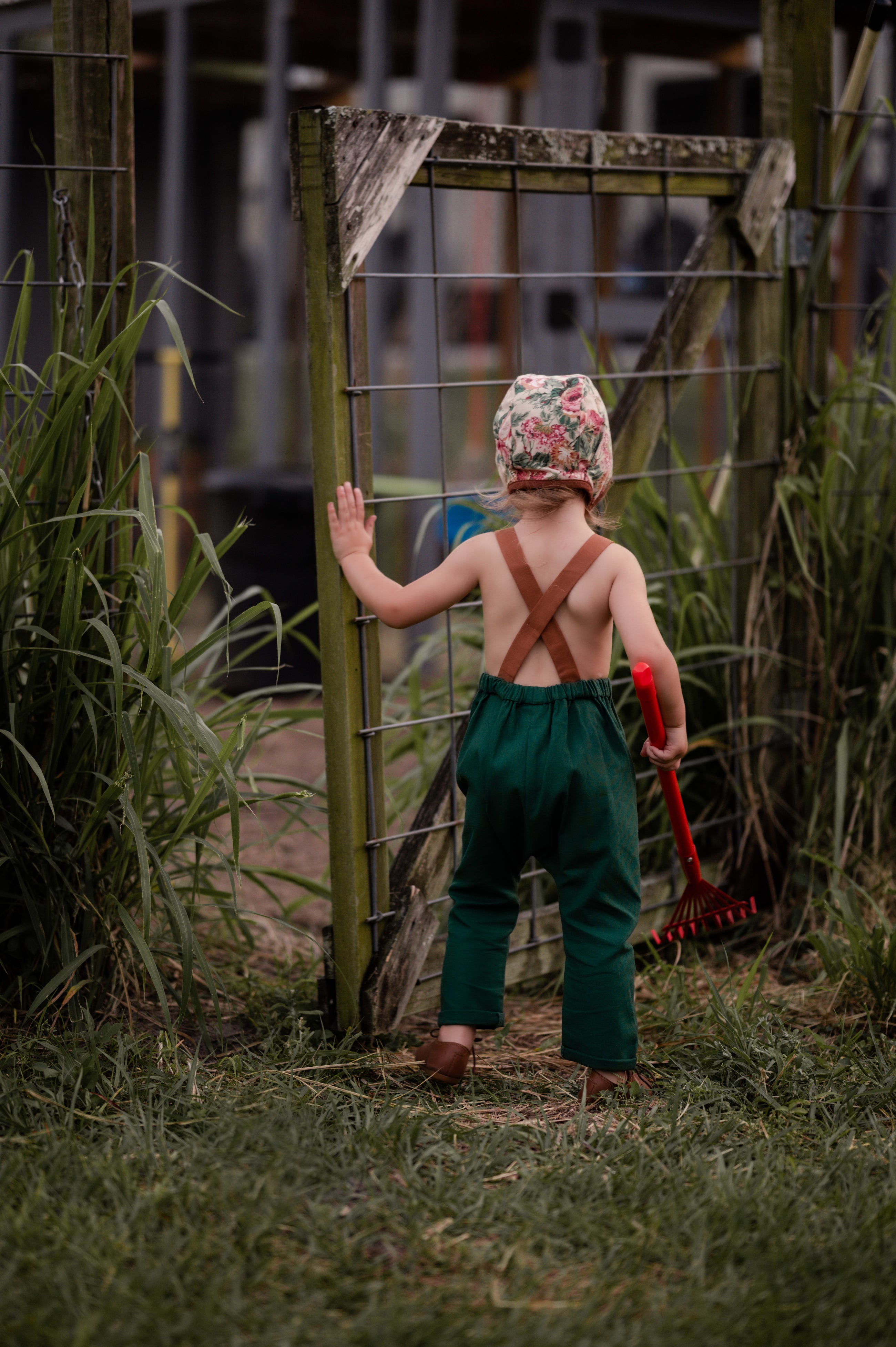 Back view of a 3-year-old girl closing a wooden gate in evergreen linen long overalls, highlighting the adjustable straps and overall functionality for everyday wear.