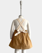 A child's short skirt displayed on a white mannequin from the back, featuring a mustard yellow fabric body with cream-colored crisscross shoulder straps and a matching fabric bow at the waist.