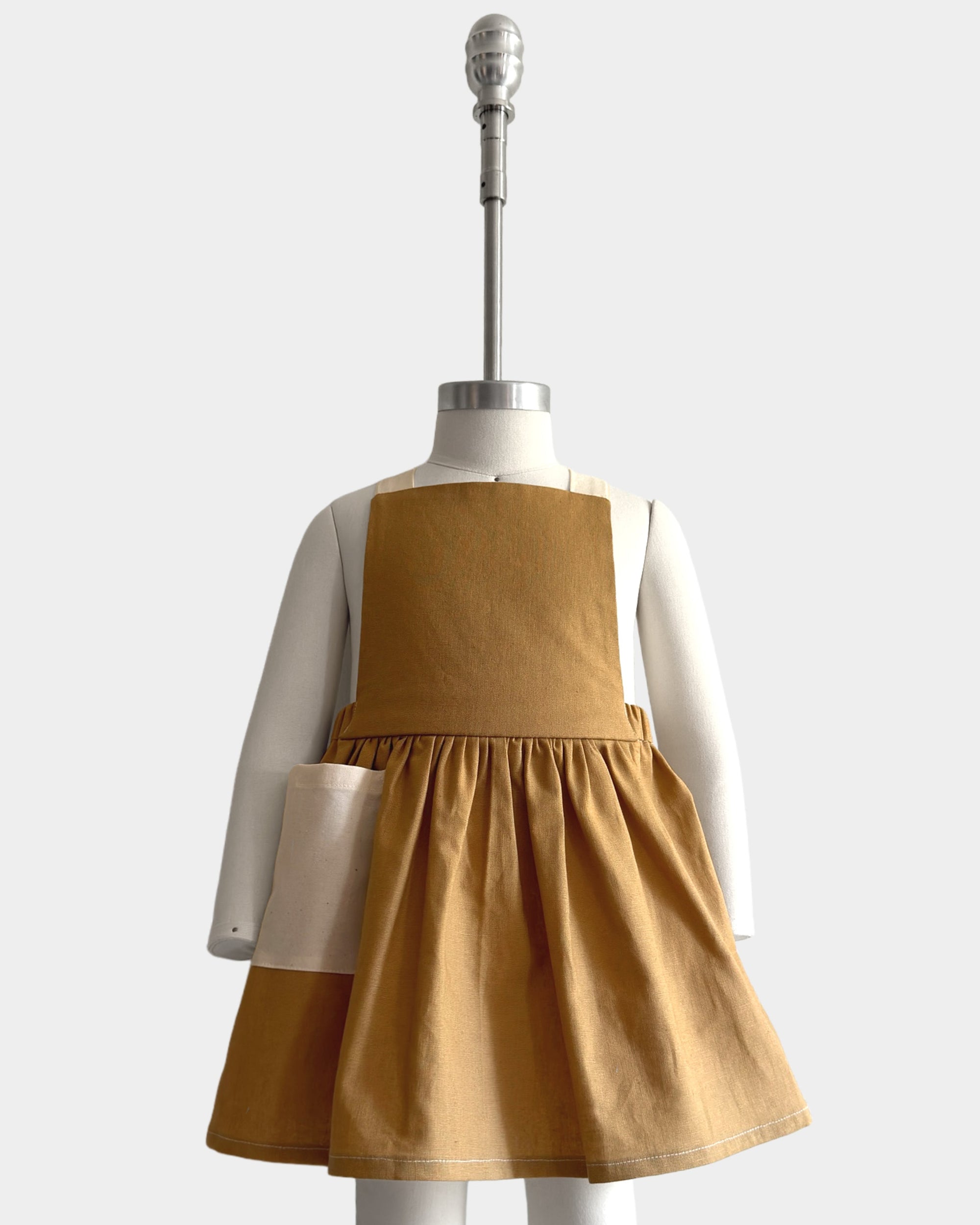 A toddler girl's sleeveless pinafore dress in a warm camel brown color, styled on a white mannequin with a white long-sleeve shirt underneath. The dress features a gathered waist and a flared skirt, photographed against a solid light gray background.