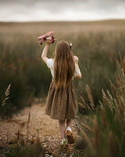 A young girl with long light brown hair is seen from behind, walking on a dirt path through a grassy field while holding a pinkish-red skateboard above her head. She is wearing a white short-sleeve top and a brown and beige checkered skirt.