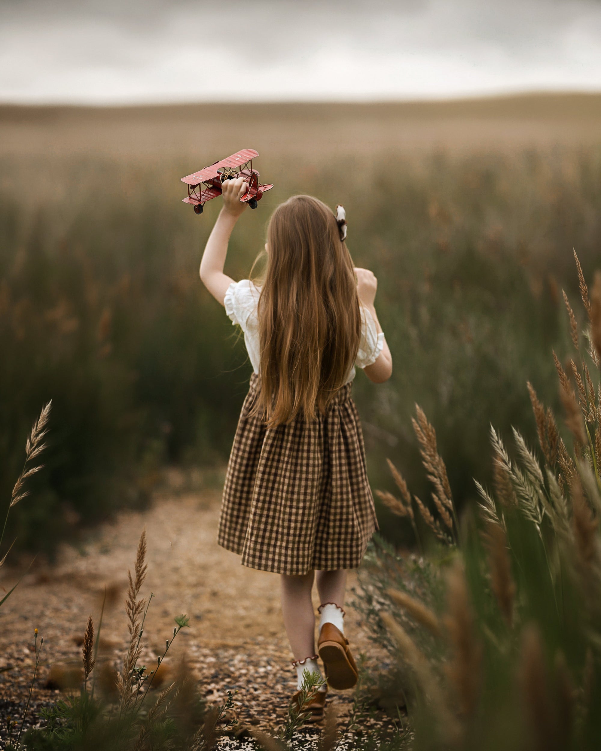 A young girl with long light brown hair is seen from behind, walking on a dirt path through a grassy field while holding a pinkish-red skateboard above her head. She is wearing a white short-sleeve top and a brown and beige checkered skirt.