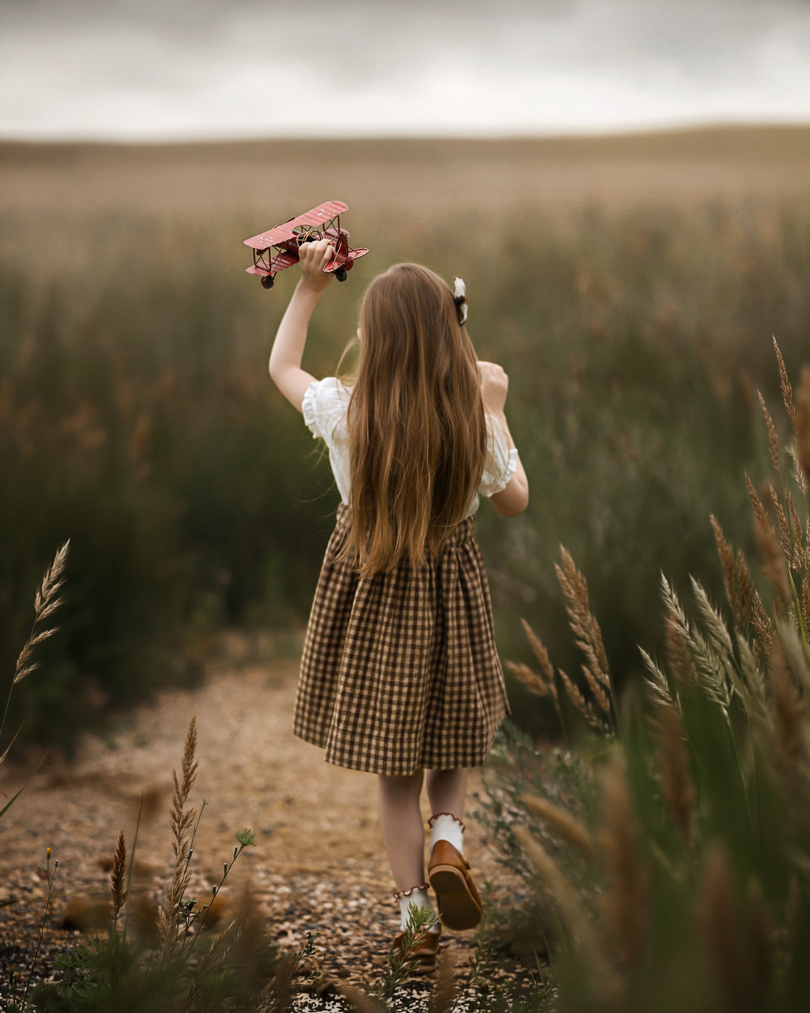 A young girl with long light brown hair is seen from behind, walking on a dirt path through a grassy field while holding a pinkish-red skateboard above her head. She is wearing a white short-sleeve top and a brown and beige checkered skirt.