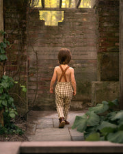 5-year-old boy wearing beige gingham linen long overalls walking away from the camera, showcasing the back view of the overalls in a playful outdoor setting. Overalls with brown contrasting straps.