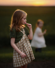 Young girl in a checkered dress standing in a field with a blurred figure in the background during sunset.