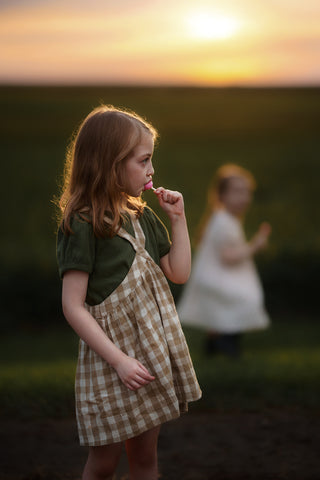 Young girl in a checkered dress standing in a field with a blurred figure in the background during sunset.