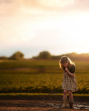 Young girl standing in a field at sunset, splashing in a puddle with rainbows eating a lollipop dressed in a checkered pinafore