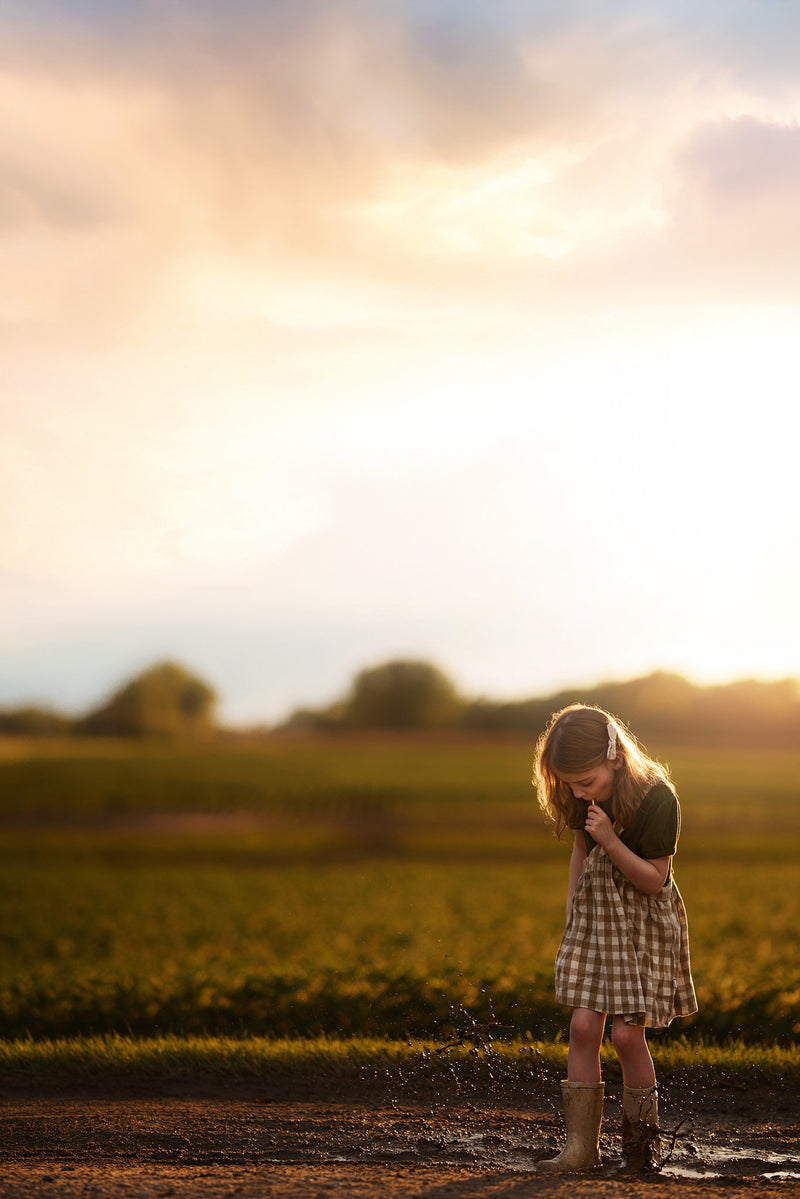 Young girl standing in a field at sunset, splashing in a puddle with rainbows eating a lollipop dressed in a checkered pinafore