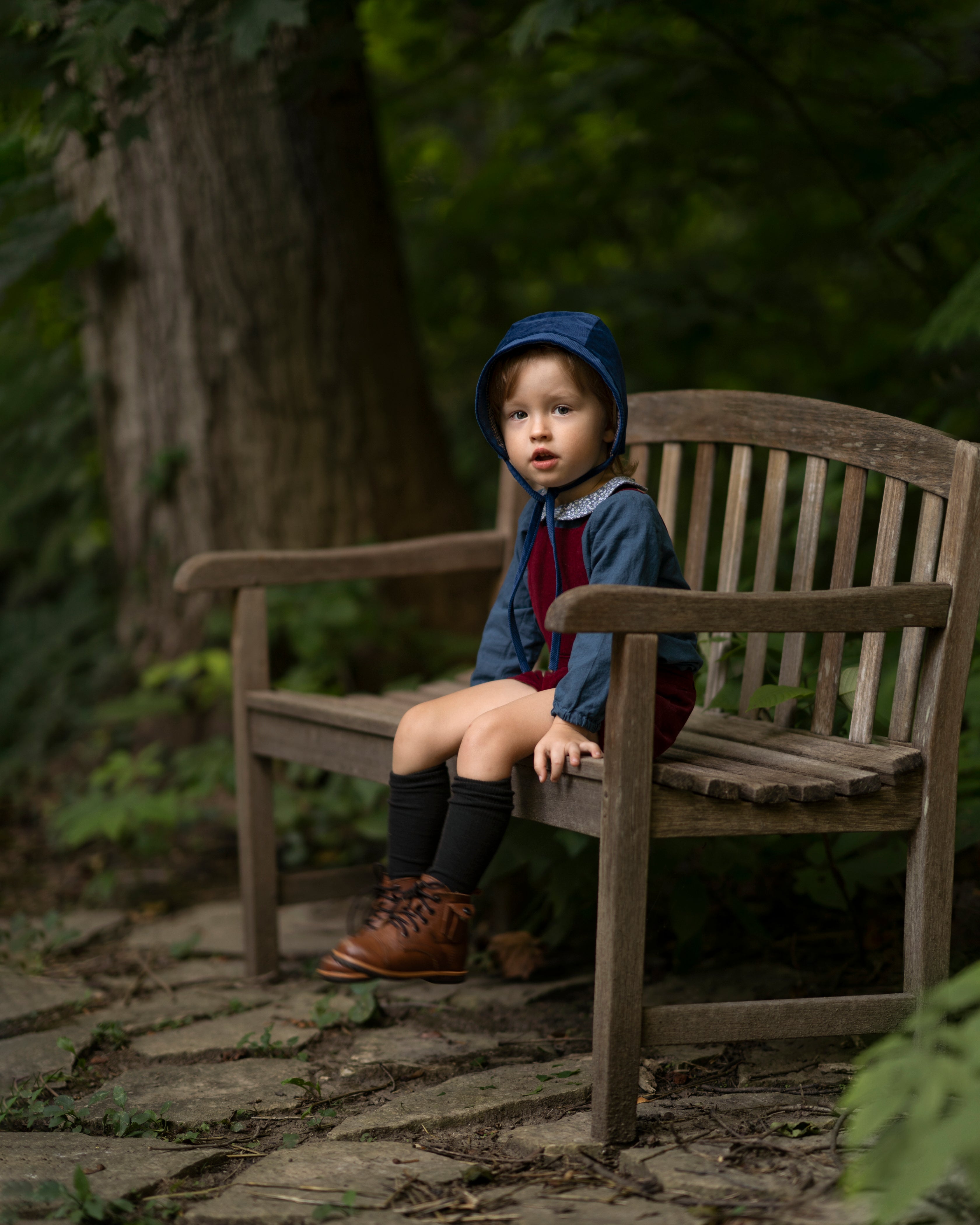 A 4-year-old boy sitting on a bench while wearing the cornflower indigo blue corduroy bonnet, highlighting its timeless accessory appeal and comfort for everyday wear.