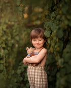 5-year-old boy wearing beige gingham linen long overalls while holding a teddy bear, featuring a gender-neutral design, breathable fabric for comfort, and adjustable straps for a perfect fit.