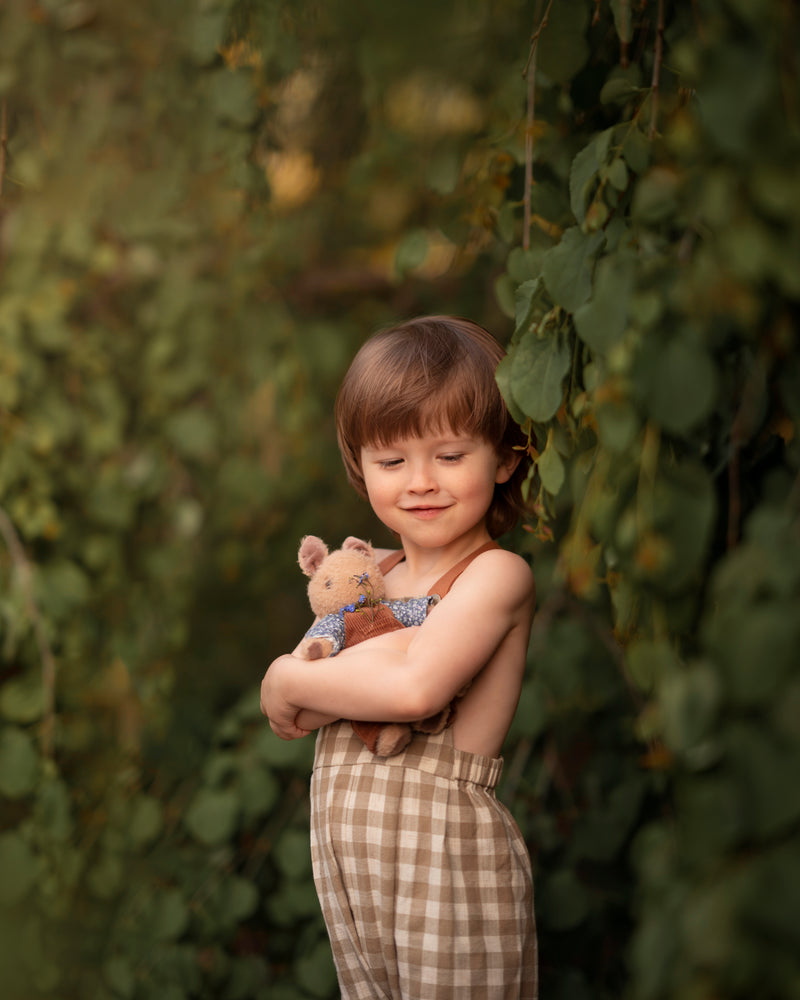 5-year-old boy wearing beige gingham linen long overalls while holding a teddy bear, featuring a gender-neutral design, breathable fabric for comfort, and adjustable straps for a perfect fit.