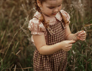 A young girl with braided hair stands in a grassy field, wearing a pink ruffled blouse and a brown gingham pinafore dress.