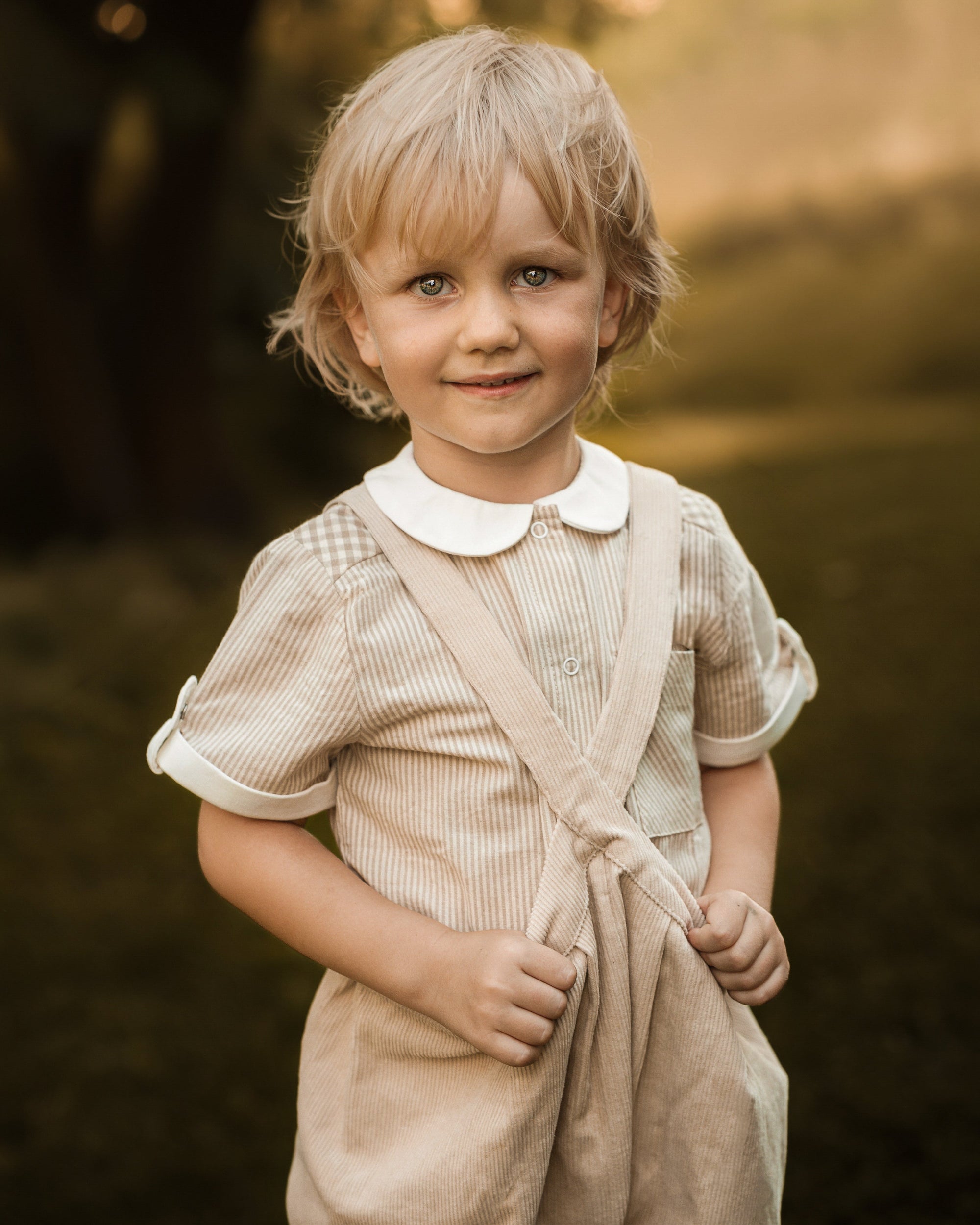 A young child with blonde hair wearing a beige ribbed pinafore-style dress with a white Peter Pan collar, standing outdoors in a warm, sunlit setting.