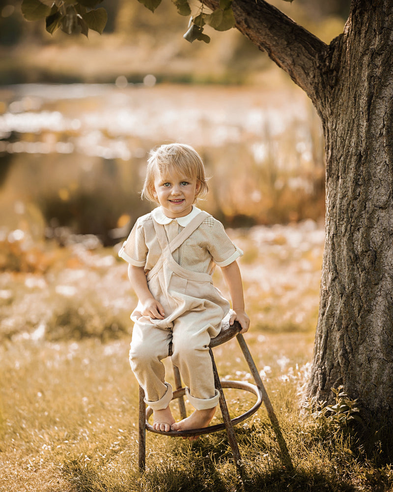 A young child wearing a cream-colored short-sleeve romper and matching pants sits on a metal stool outdoors next to a tree, with a blurred natural background.
