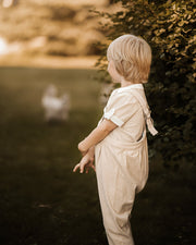 A young child with short blond hair stands outdoors in profile near a leafy bush, wearing a light cream or off-white short-sleeve jumpsuit or romper. The background is softly blurred with warm golden light, grass, and trees.