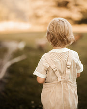 A young child seen from behind, wearing a cream-colored ribbed short-sleeve shirt with matching cream overalls featuring adjustable shoulder straps, standing in an outdoor setting with a warm, golden, blurred background.