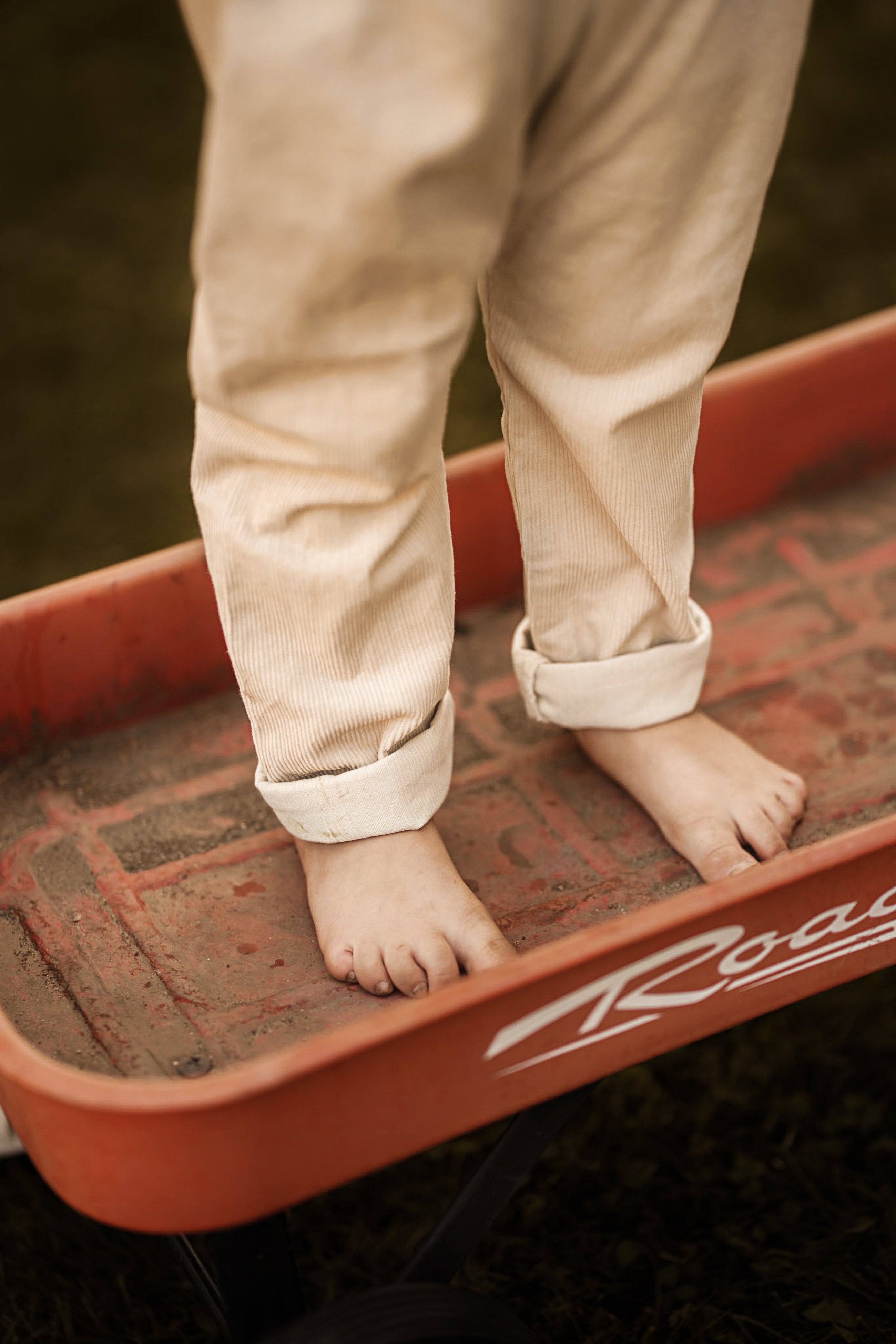 A close-up lifestyle shot of a child's bare feet standing in a red wagon while wearing beige corduroy pants.