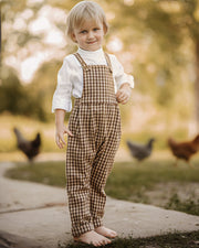 A toddler stands barefoot outdoors on a path, wearing brown and beige gingham bib overalls over a white long-sleeve shirt. Chickens are blurred in the background among trees.
