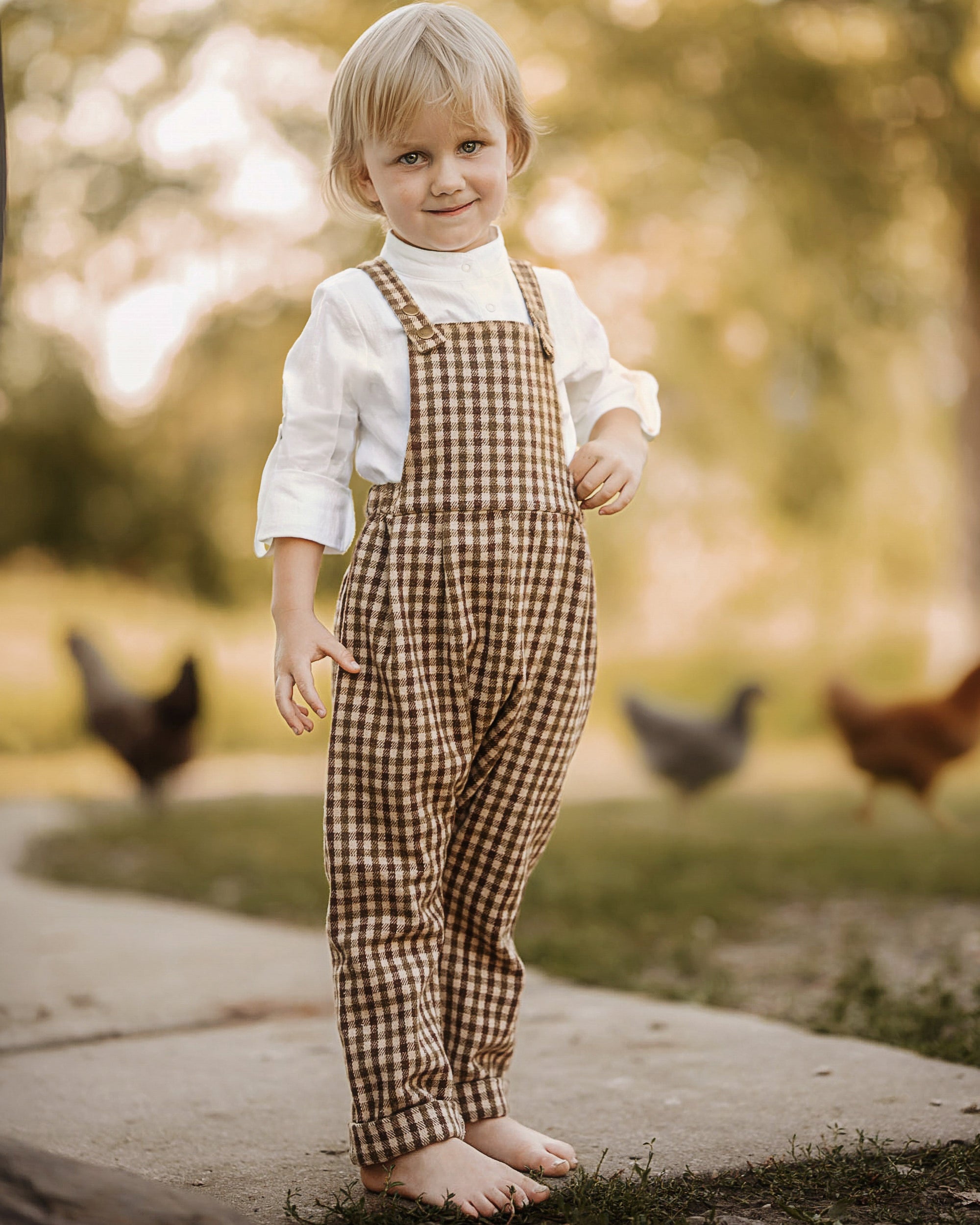 A toddler stands barefoot outdoors on a path, wearing brown and beige gingham bib overalls over a white long-sleeve shirt. Chickens are blurred in the background among trees.