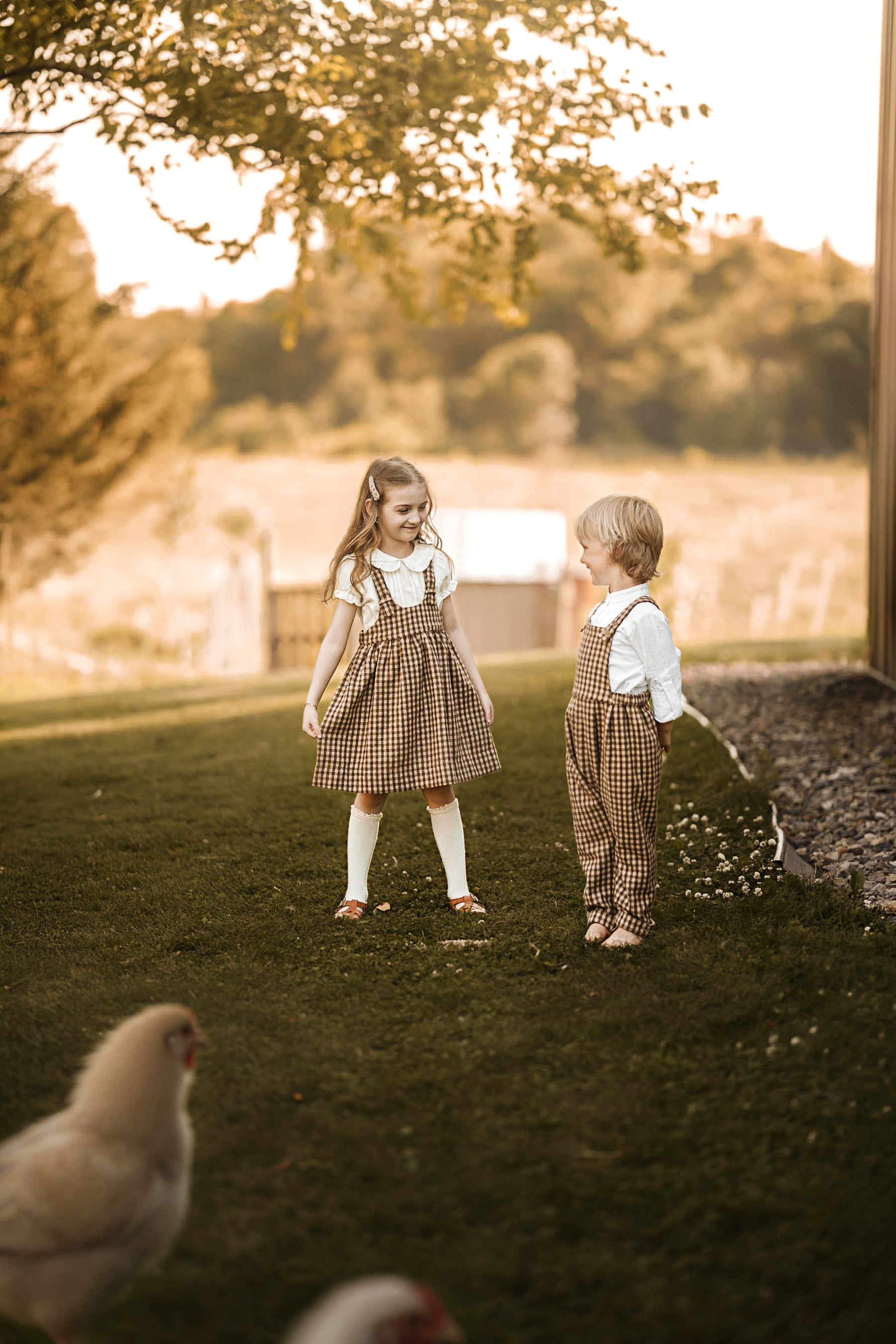 A young girl and a toddler stand on a grassy lawn outdoors, wearing matching brown and beige gingham check puffer dresses and overalls with white shirts. A chicken is visible in the blurred foreground, and trees and a warm-toned outdoor setting fill the background.