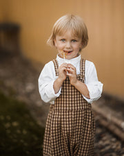A young blonde boy wearing a white collared shirt and brown gingham overalls, holding a lollipop in his mouth and smiling, set against a warm, blurred background.