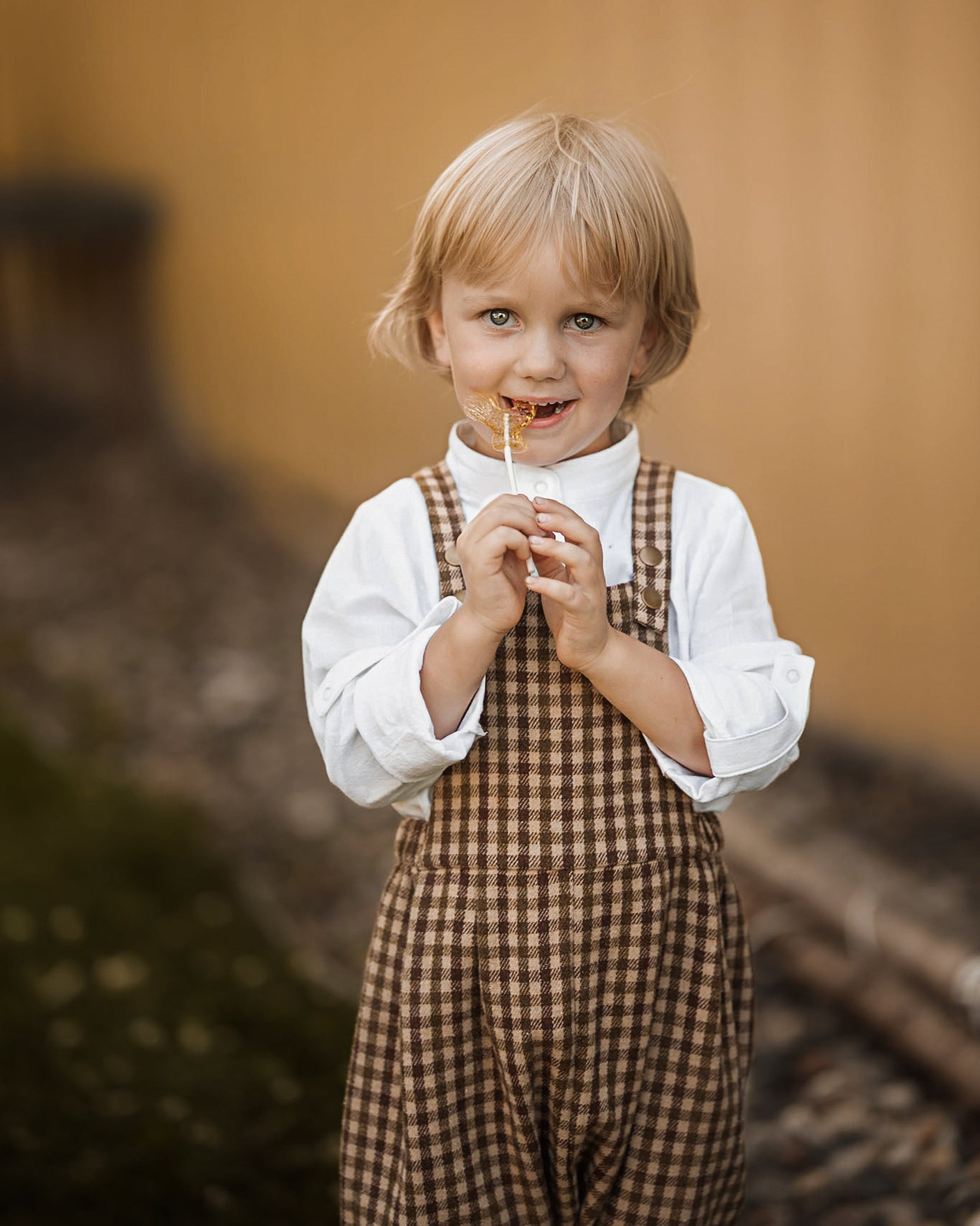 A young blonde boy wearing a white collared shirt and brown gingham overalls, holding a lollipop in his mouth and smiling, set against a warm, blurred background.