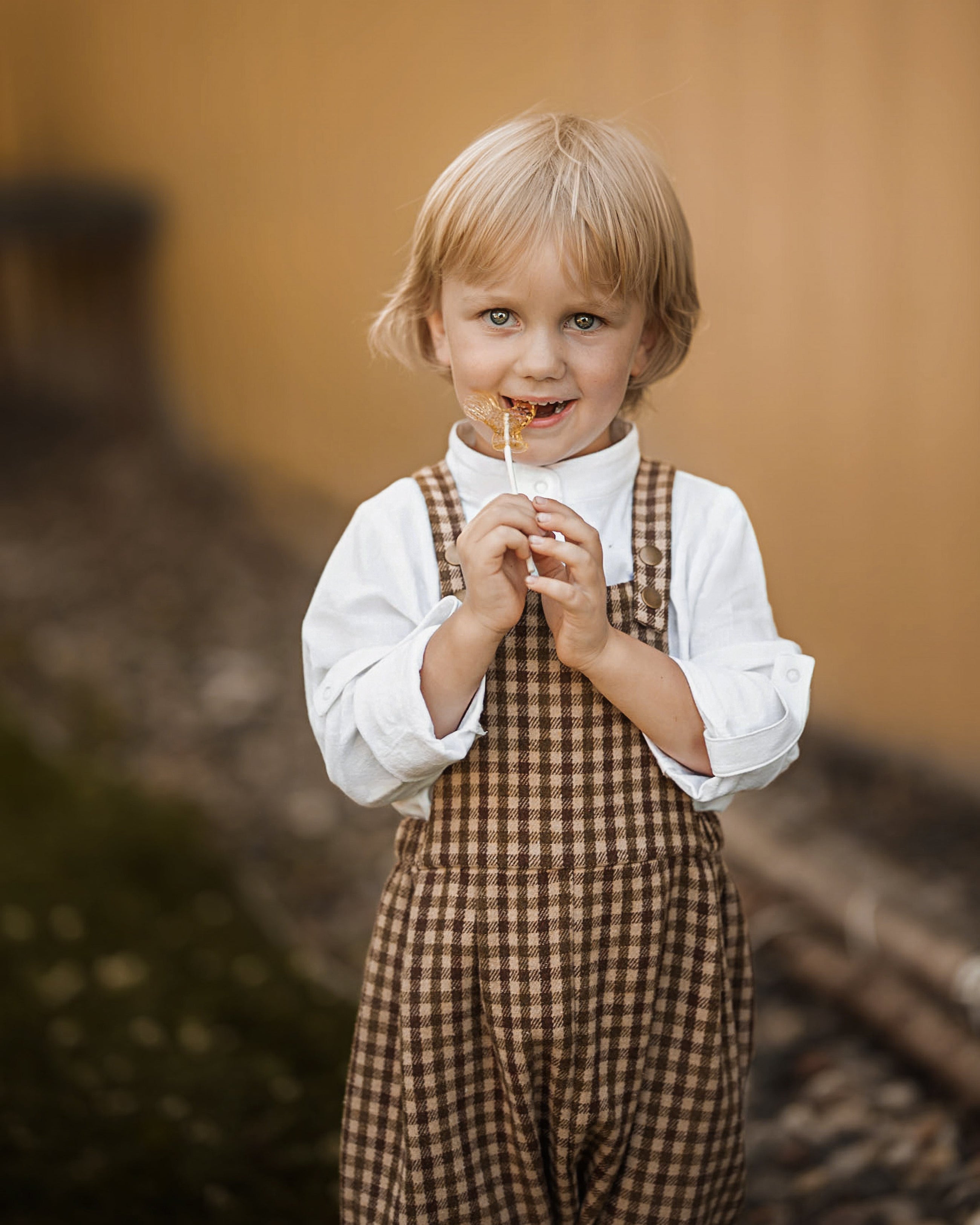 A young blonde boy wearing a white collared shirt and brown gingham overalls, holding a lollipop in his mouth and smiling, set against a warm, blurred background.