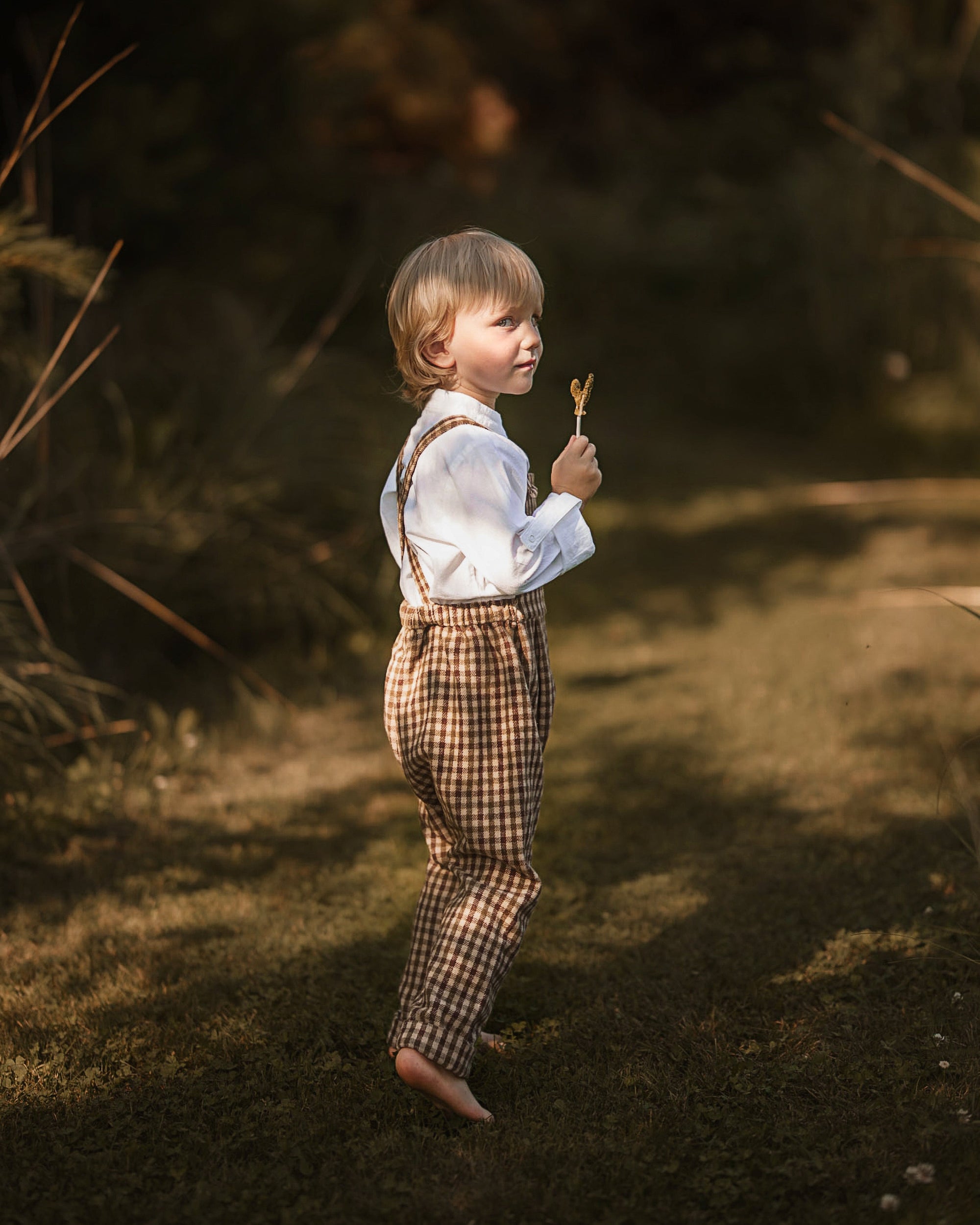 A young child standing outdoors in a wooded setting, wearing a white long-sleeve shirt and brown-and-beige gingham suspenders, holding a small object in one hand.
