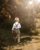 A young child seen from behind walking on a sunlit grassy path outdoors, wearing a white shirt, brown suspenders, and brown-and-white patterned trousers.