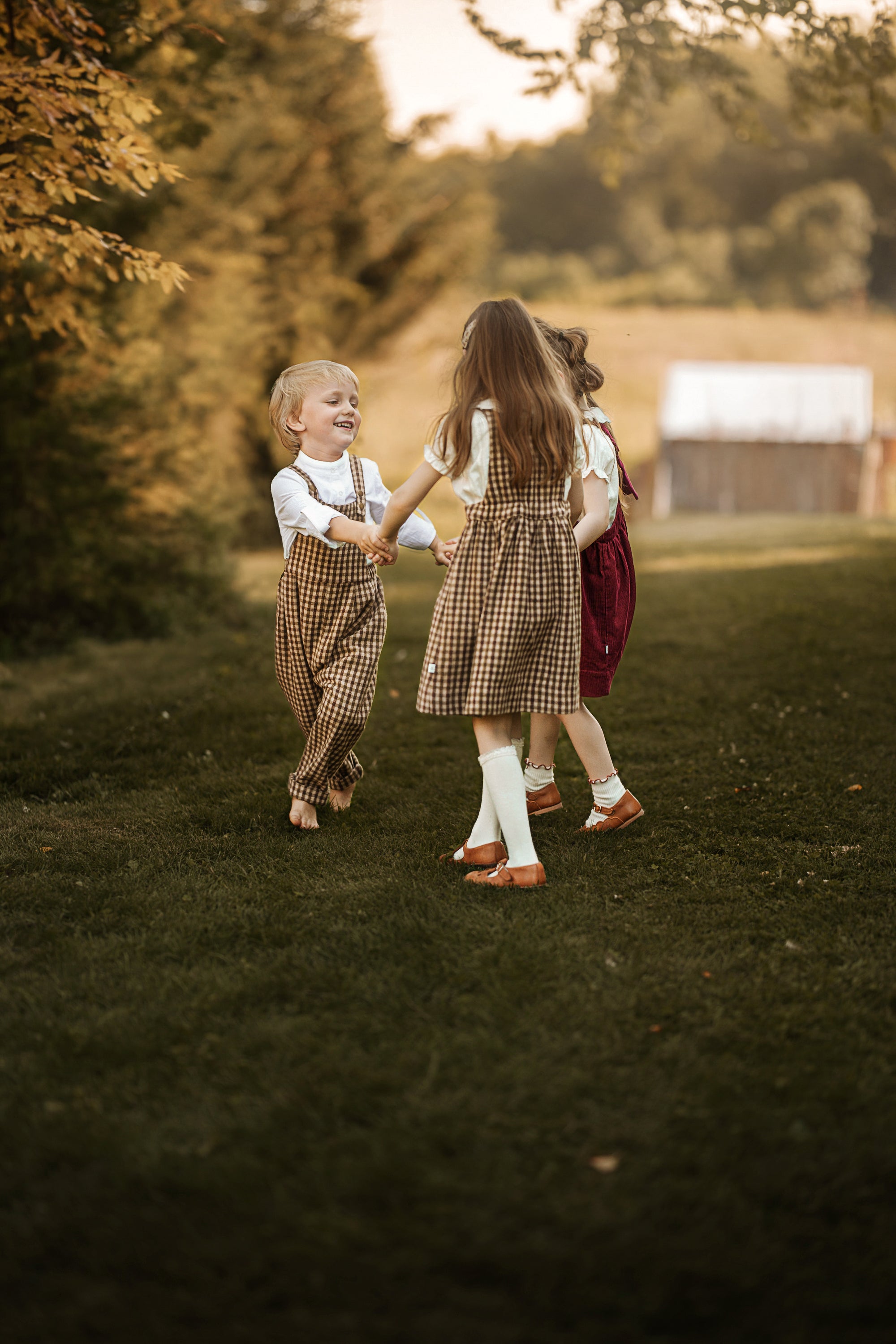 Three children playing outdoors on a grassy lawn in warm golden hour light, wearing matching brown plaid pinafore-style dresses and overalls with white shirts and brown shoes.