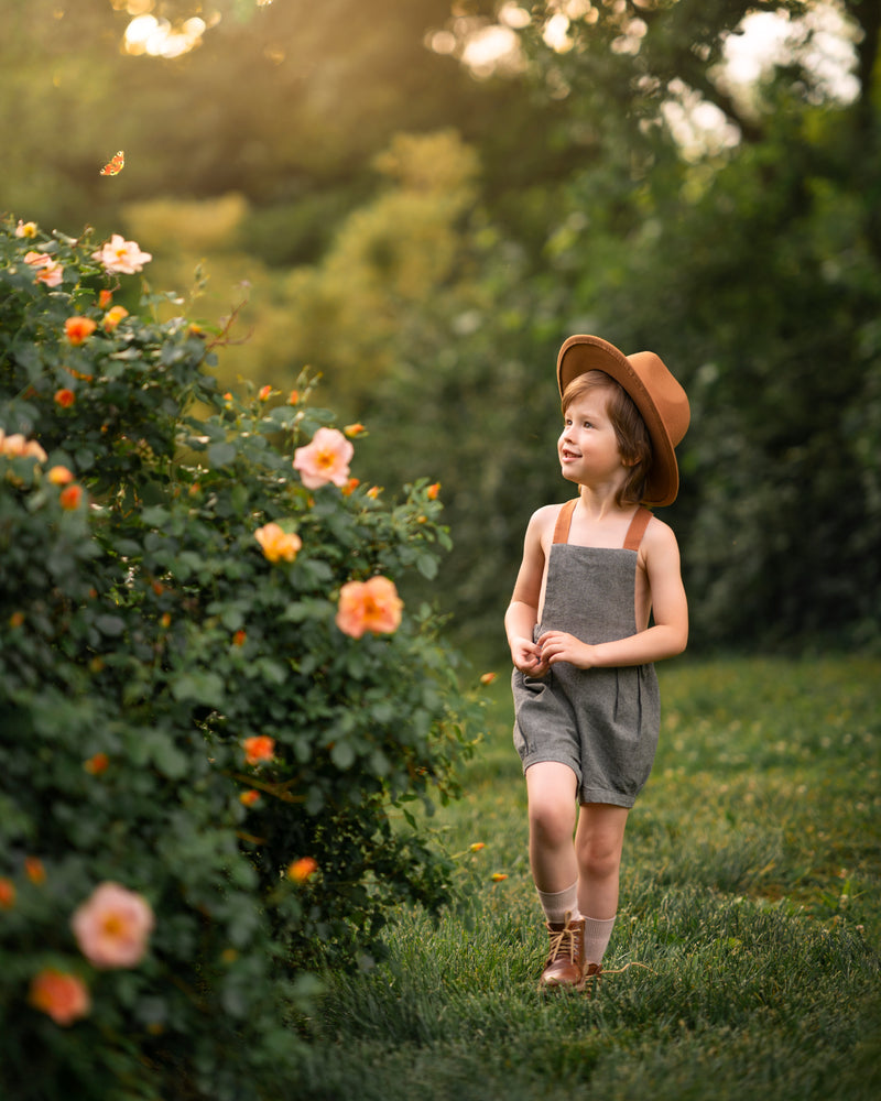 Little boy in green overalls and a hat walking near a rose bush looking at a butterfly flying