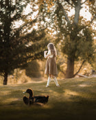 A young girl stands on a grassy hill in a sunlit park, viewed from behind as she looks toward trees and ducks in the foreground. The scene is warm and golden with soft-focus foliage and no clear retail product isolated.