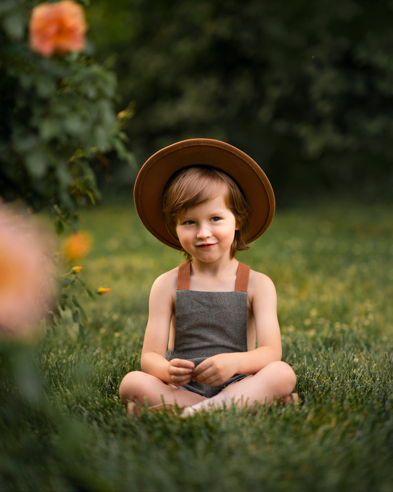 Little boy in green overalls and a hat sitting with cross legs on grass