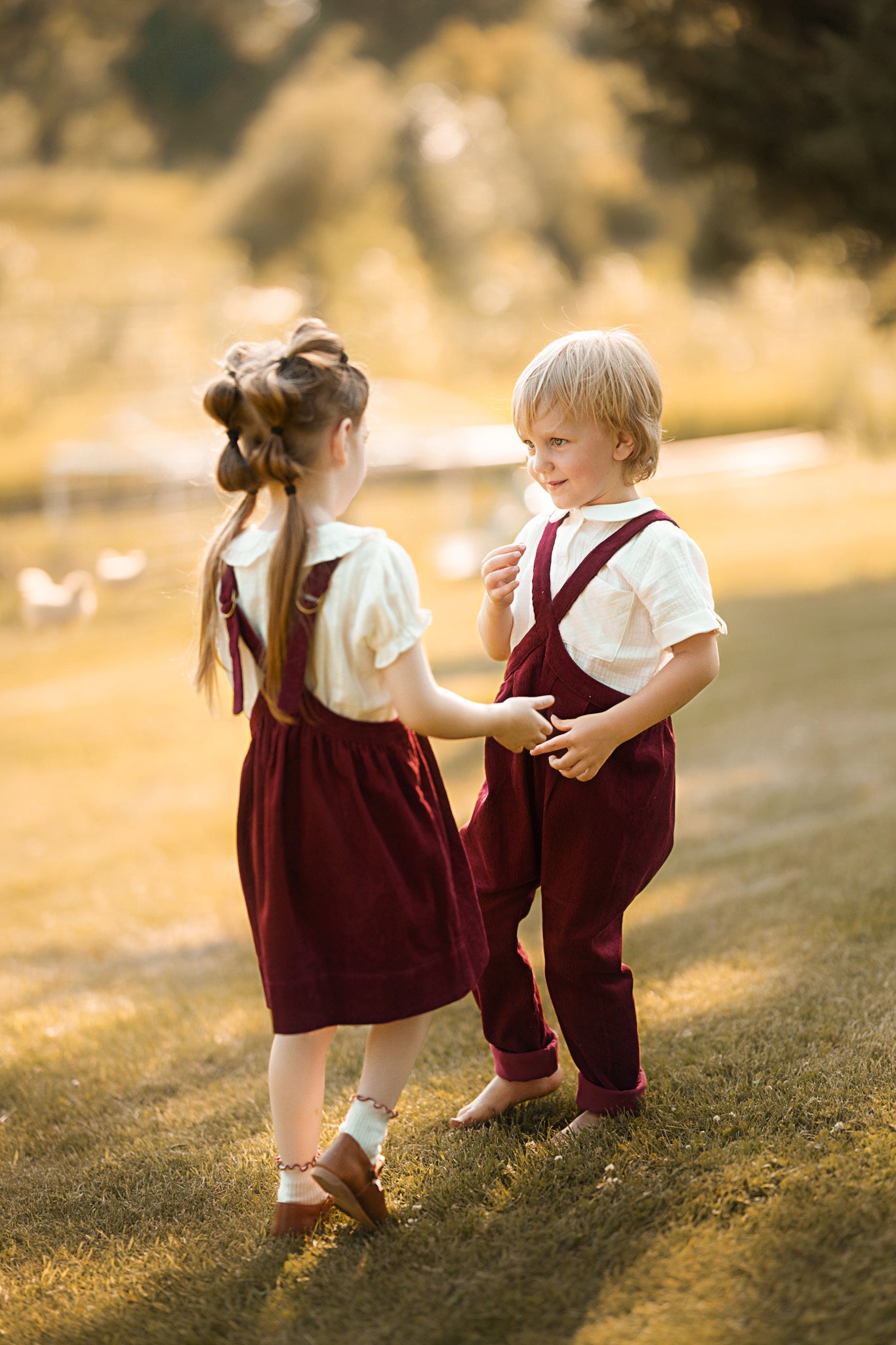 Two young children standing on grass in a sunlit outdoor setting, wearing matching outfits with white short-sleeve shirts and dark red pinafore-style overalls. The girl has her hair in two buns, and the boy has light blond hair.