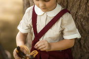 A young child wearing a cream and white striped short-sleeve shirt with a white collar and dark red corduroy suspenders, standing outdoors next to a tree trunk.