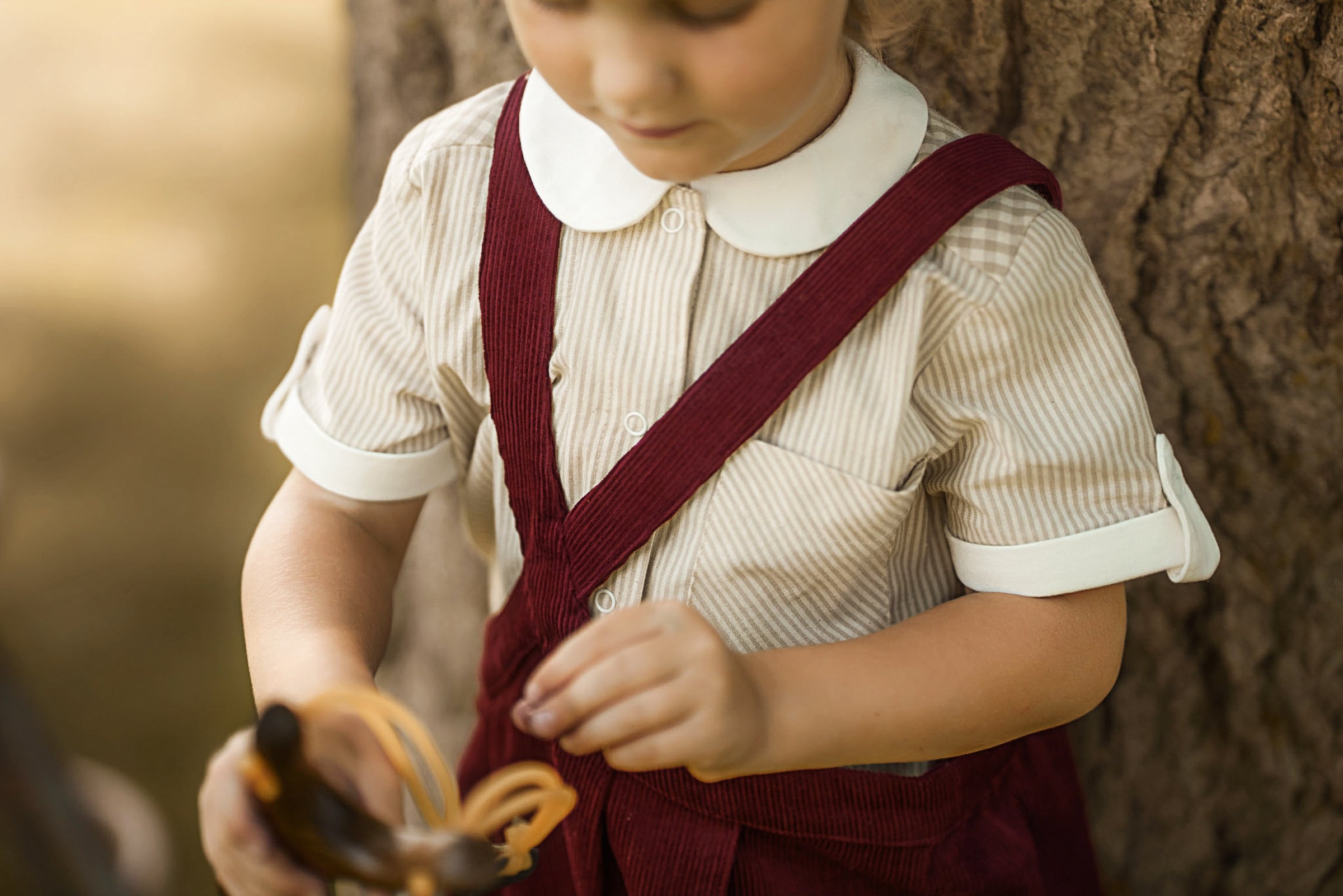 A young child wearing a cream and white striped short-sleeve shirt with a white collar and dark red corduroy suspenders, standing outdoors next to a tree trunk.