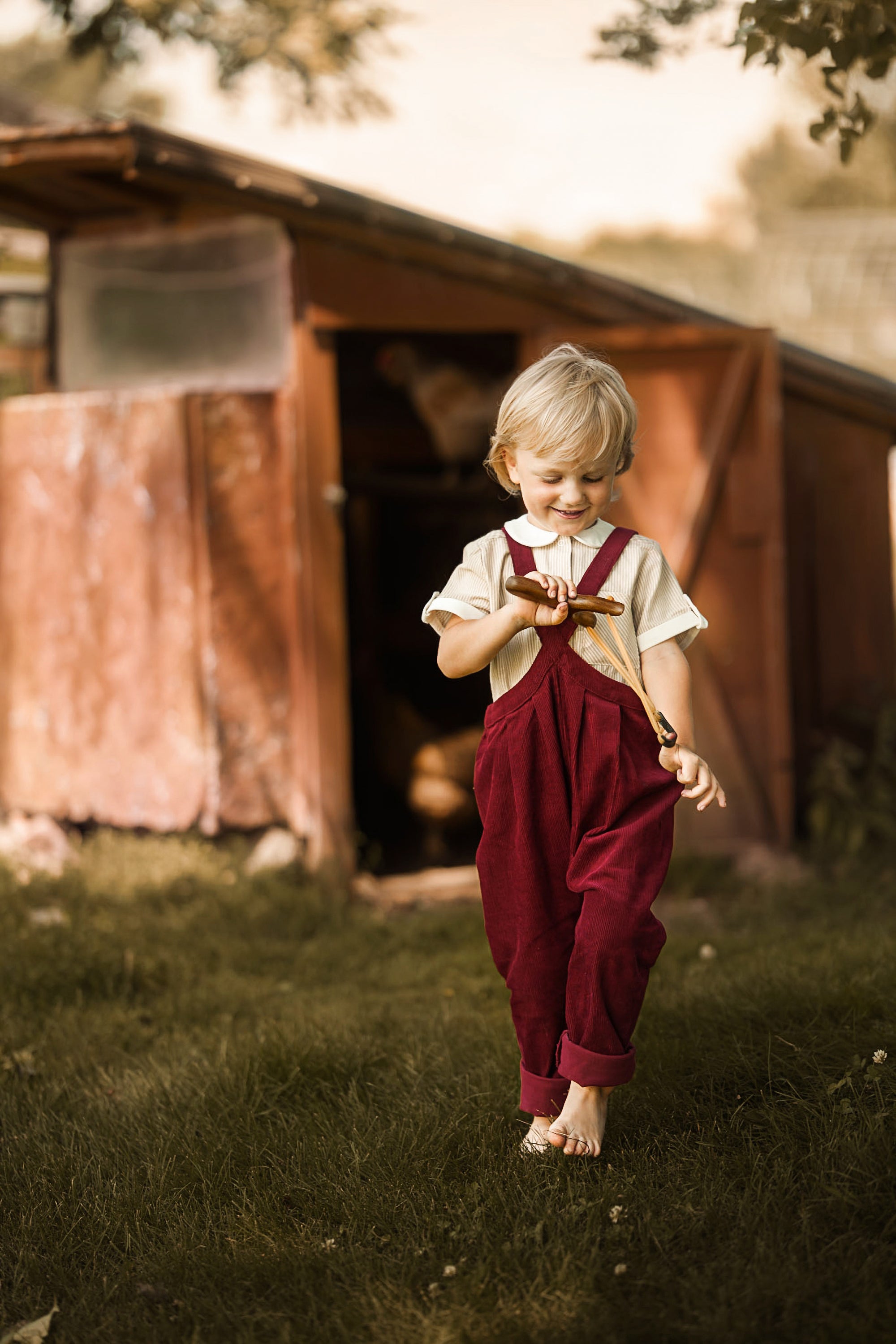 A young blonde child walking barefoot in a grassy yard in front of a rustic wooden shed, wearing a white short-sleeve shirt and dark red corduroy overalls.