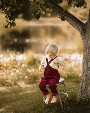 A young child with blonde hair sits barefoot on a small stool in a grassy outdoor setting under a tree, wearing a cream shirt and dark red overalls.