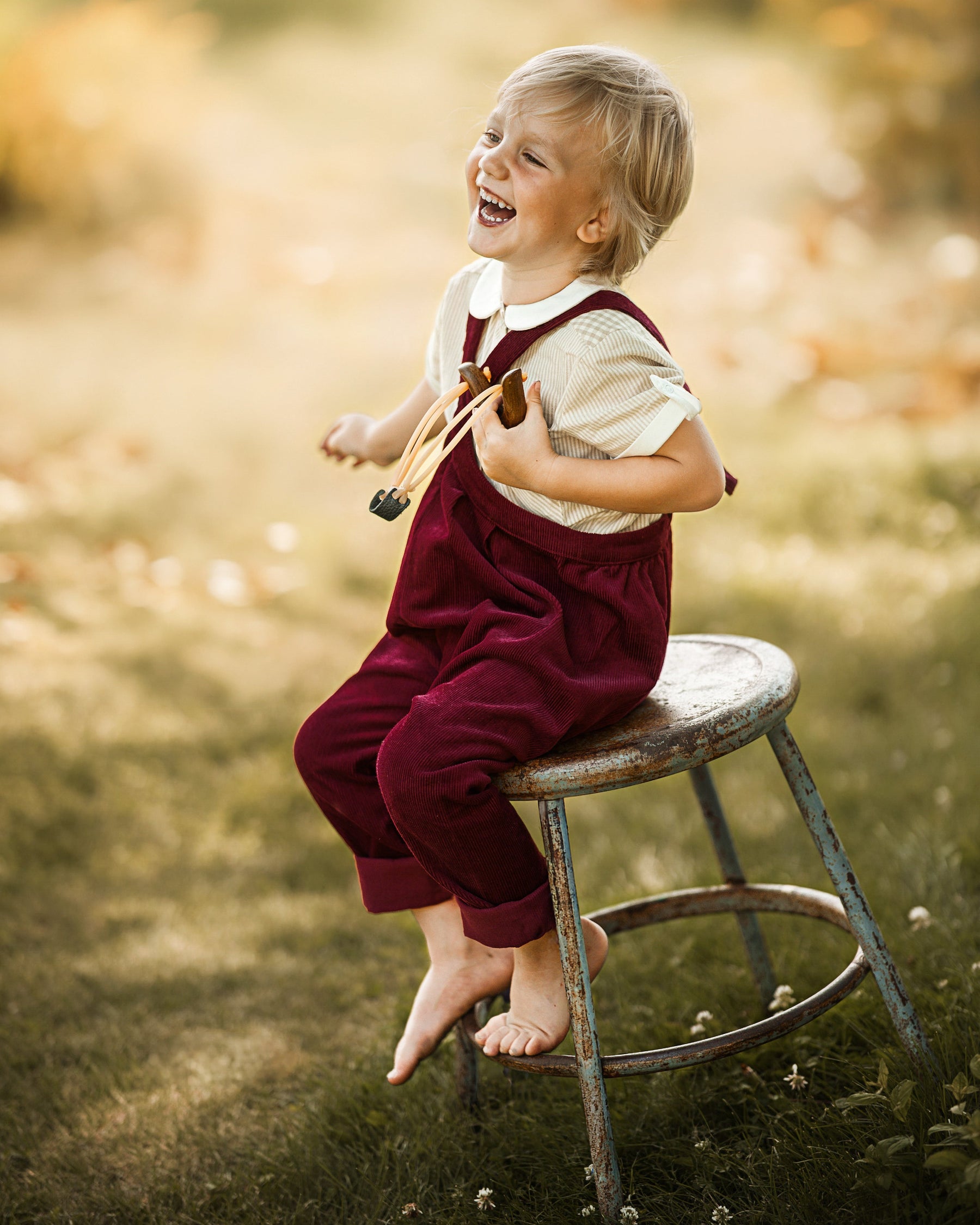 A young child with blonde hair sits barefoot on a rustic metal stool outdoors, laughing and looking upward. The child is wearing burgundy overalls over a cream short-sleeve shirt.