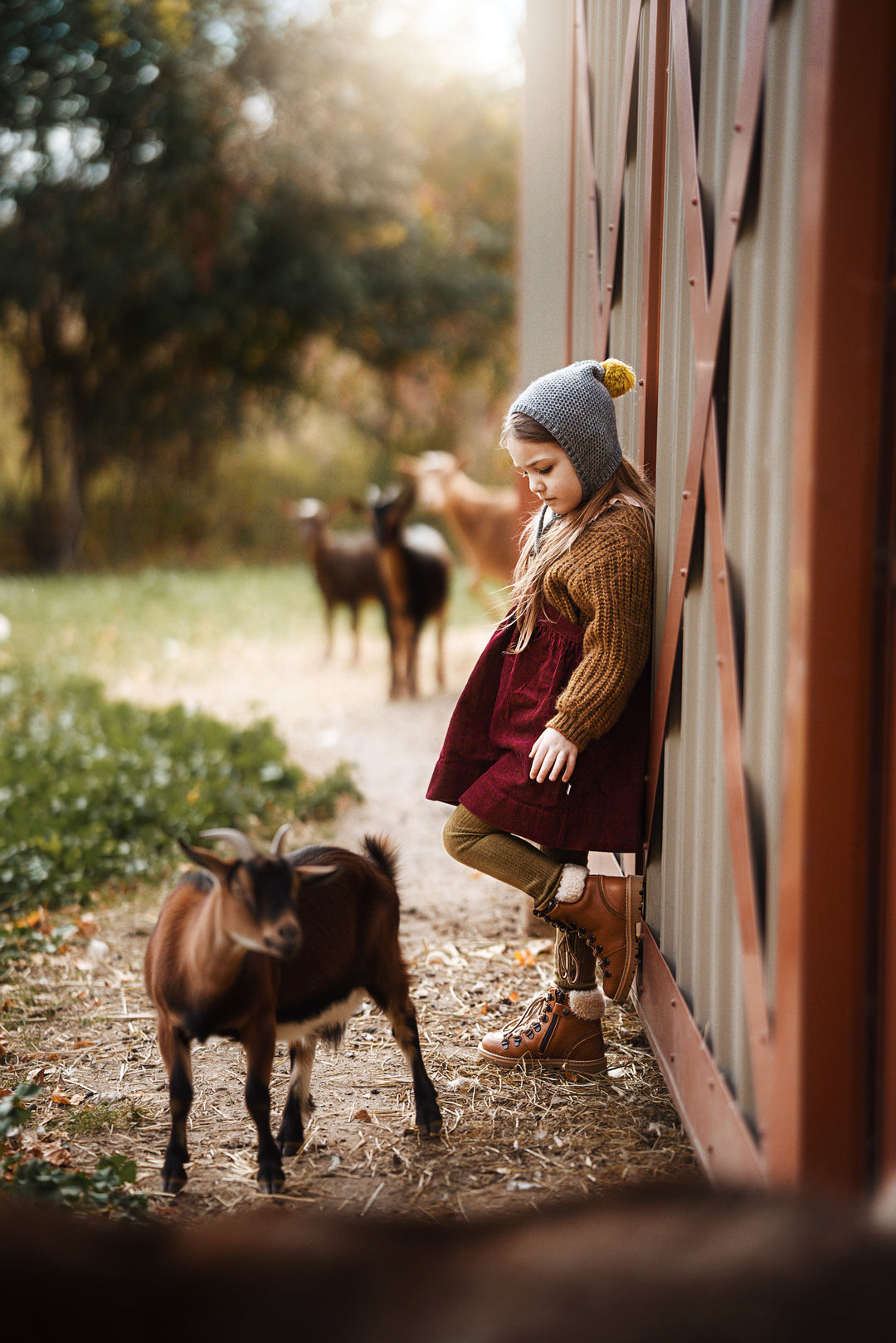 A young girl in a cozy knit hat, brown sweater, and burgundy dress leans against a red barn door while a small brown goat stands nearby on a dirt path. Other goats are visible in the soft-focus background outdoors.