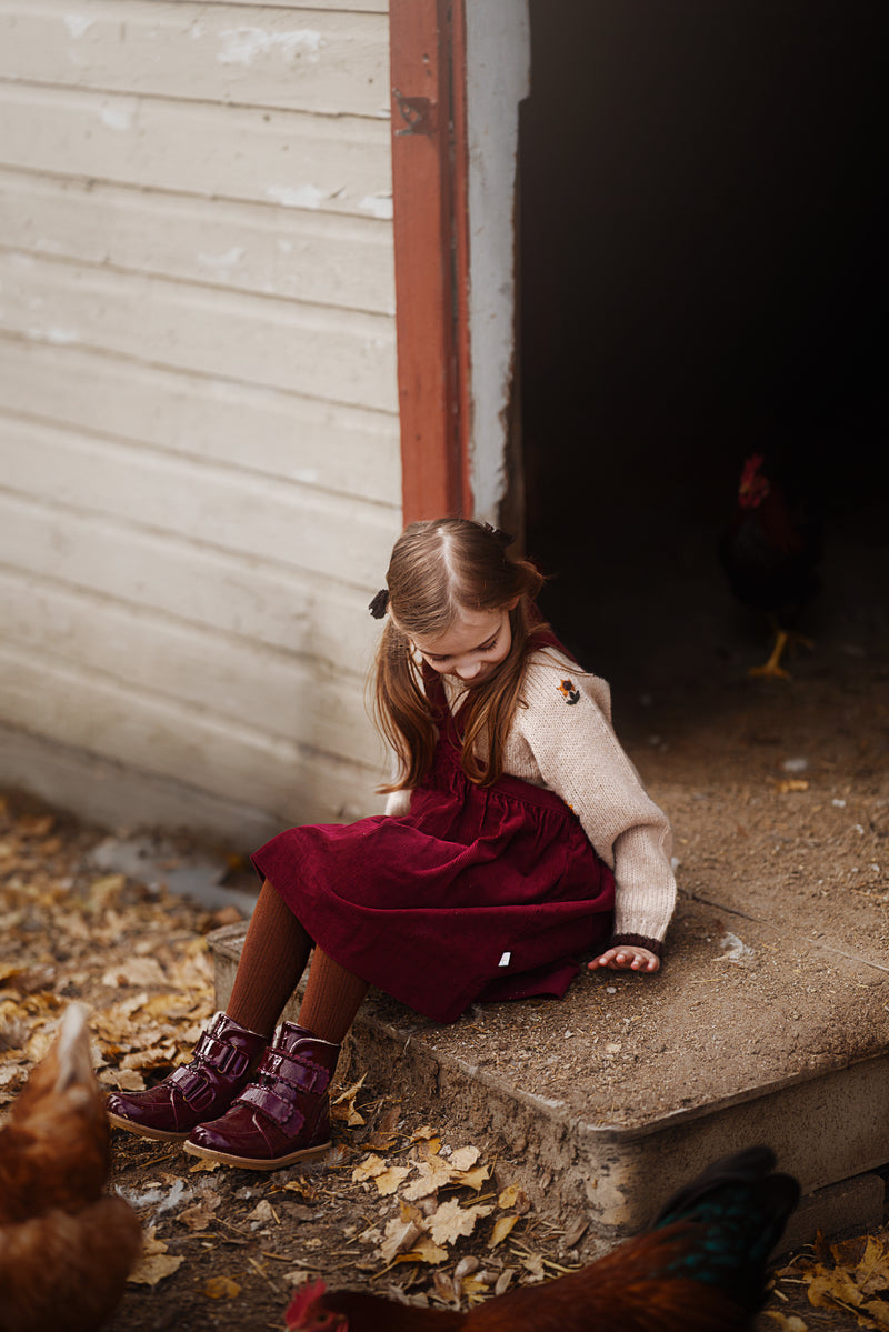 A young girl sits on concrete steps in front of a white wooden barn door, wearing a cream knit sweater, a burgundy velvet dress, and dark burgundy boots. Chickens are visible on the ground and in the barn doorway.